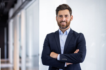 Naklejka premium Portrait of a smiling young successful male businessman standing in the office in a suit and looking at the camera with his arms crossed on his chest
