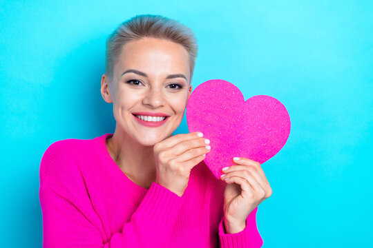 Photo of cute positive woman with bob hairdo dressed pink sweater hold heart near face on valentine day isolated on teal color background