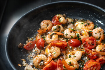 Shrimps with tails are sauteed with tomatoes, herbs and garlic in olive oil in a black frying pan, cooking a Mediterranean seafood meal, selected focus - 700375779