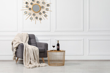 Comfortable armchair, blanket and wine on side table near white wall, space for text. Interior...