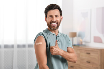 Man with sticking plaster on arm after vaccination showing thumbs up at home