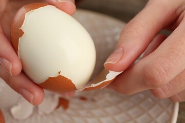 Woman peeling boiled egg over plate, closeup