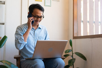 The young man asian is working from home in the living room, using his mobile phone and computer to command his work remotely.