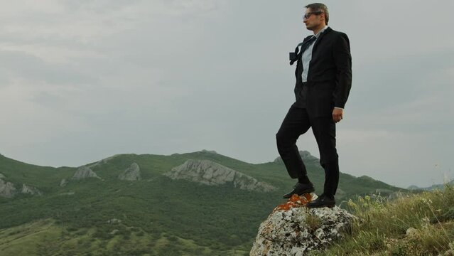 An adult man in a business suit fluttering in the wind stands on a stone against the backdrop of a mountain range and adjusts his glasses. The concept of stress resistance and psychological stability