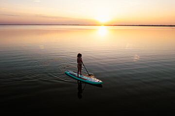 A woman in mohawk shorts stands on a SUP board at sunset in a lake against a pink-blue sky and...