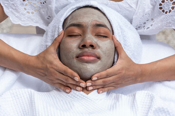 Young African American black woman lies down while a facialist uses facial cream and mask to...