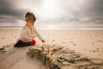 Girl with blond hair builds sand castle on beach with gray clouds