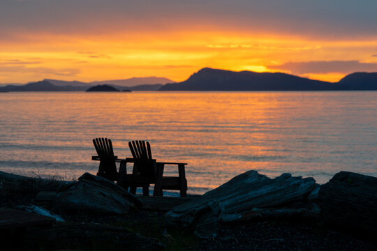 Soft focus image of two chairs on a beach outlined against the ocean and mountains under a dramatic orange post-sunset on Orcas Island, Washington