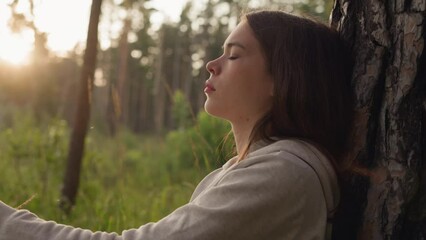 Woman thinks with closed eyes leaning on tree trunk at sunset. Depressed lady considering psychology help to cope with stress in calm atmosphere of evening in nature