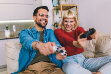 Boyfriend and girlfriend playing video game with joysticks in living room. Loving couple are...