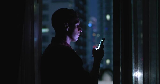 Man standing at window of skyscraper late in evening with phone in hand. Millennial reads messages on phone after long day at work. Cinematic shot of handsome man in light of phone and city lights.