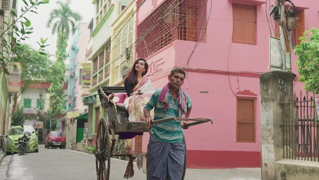 An aged or elderly Indian Asian male human or hand pulled rickshaw puller pulling a rickshaw with a young traditional woman or female is sitting in an ethnic saree in a narrow lane with old buildings