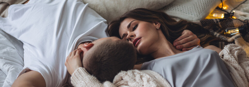 Banner relationships. Beautiful happy loving young couple relaxing in bed, looking at each other. Cozy home atmosphere, tenderness, closeness. Embracing, kissing. Lazy weekend, slow living concept