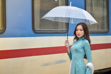 Gorgeous girl wearing a jade green ao dai traditional costume of Vietnam, waiting at the rail station with transparent umbrella