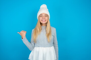 Lovely Caucasian blonde girl wearing blue sweater and wool beanie pointing aside with forefinger, showing at copy space having news about bargains