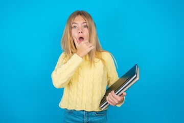 Naklejka premium Caucasian blonde girl wearing yellow sweater holding notebooks Looking fascinated with disbelief, surprise and amazed expression with hands on chin