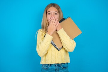 Stunned beautiful caucasian teen girl wearing yellow sweater covers both hands on mouth, afraids of...