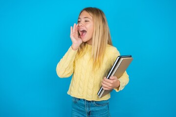 beautiful caucasian teen girl wearing yellow sweater look empty space holding hand face and screaming or calling someone.