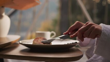 Woman cuts piece of pancake at table in glamping house closeup. Lady eats delicious breakfast by glass wall in hotel. Guest service at resort