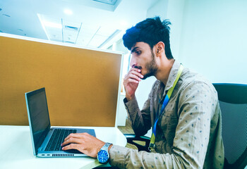 A young Indian man diligently working on his laptop on the desk.