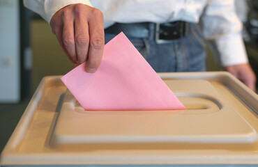 Stimmabgabe im Wahlbüro: Nahaufnahme der Hand eines Mannes, der seinen Stimmzettel in die Wahlurne...