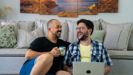 Young gay couple sit next the sofa relax together at home using computer, buying through e-commerce retail e services, watch new movie online, websurfing