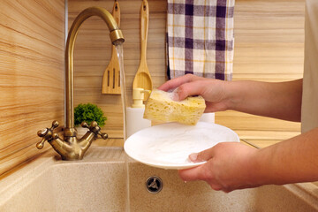 Washing dishes under the tap in the kitchen sink. Women's hands washing a white plate with a sponge...