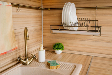 Kitchen sink in the corner with a plates, sponge, dispencer, towel and green plant. Kitchen with...