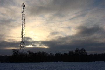 Winter morning. A tower of cellular communication against the background of the cloudy sky with a rising sun. A dark strip of the forest under a tower. Below snow-covered field. Contrast picture.