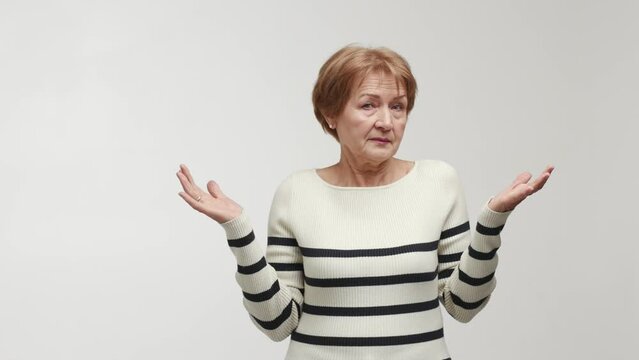 An elderly woman with a puzzled gaze shrugs, shakes a head, raises eyebrows, spreads arms and palms to the sides, shows emotions of abashment, confusion and bewilderment. Gesture of perplexity, symbol