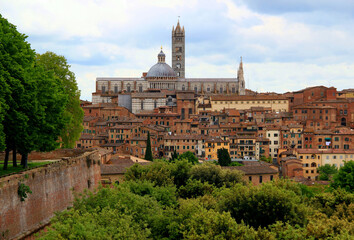 Fototapeta na wymiar Panoramic view of the historic part of the city of Siena with the Duomo di Siena and green trees and bushes in the foreground against a blue sky with clouds in the Tuscany region of Italy
