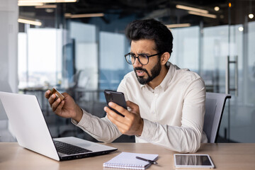 Problems with your credit card account. Worried young Indian man sitting at desk in office, holding card and phone and looking frustrated at laptop