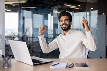 Portrait of a happy young Indian man sitting in the office and holding the phone, rejoicing in...