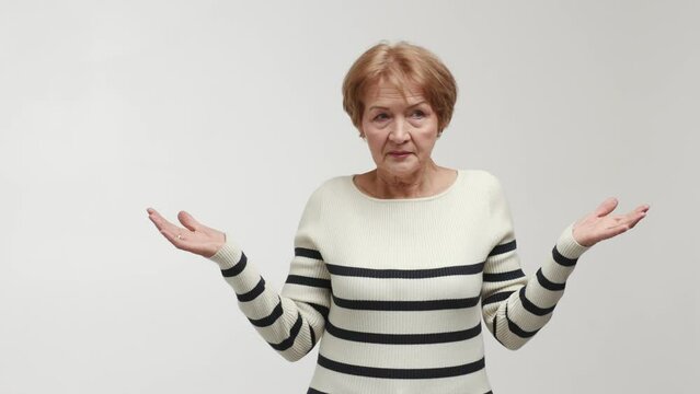 An elderly woman with a puzzled face and shocked gaze shrugs, turns a head, spreads arms and palms to the sides, shows emotions of abashment, confusion and bewilderment. Gesture of perplexity, symbol