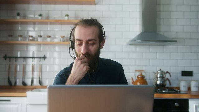 Bearded with hair bun businessman sitting in kitchen wear wireless headset having video conference calling on laptop computer talk by webcam in online chat