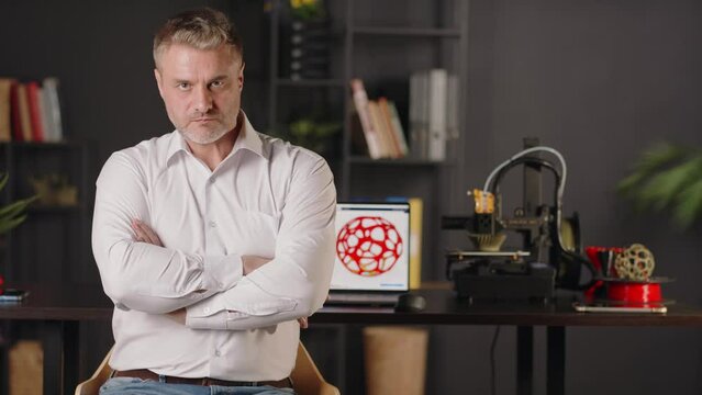 A positive smiling middle aged man graphic designer uses a laptop and creates a sketch of red three dimensional model of geometric solid. A 3d printer machine works in automated autonomous process
