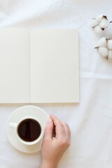 Opened note, cotton branch, hand with a coffee cup on a white sheet. Cozy composition on a bed