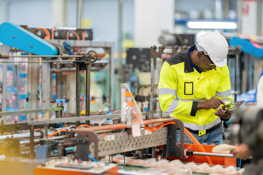 African male engineer touching and looking at laptop at work. Wearing a vest, safety helmet. Around there are working machines. It is in the plastic and steel industry of a large business company.