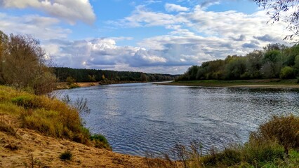 On the sandy banks of the river that makes a bend, grass grows and a forest stands. In the autumn the leaves on the trees turn yellow. There are ripples on the water. The weather is cool