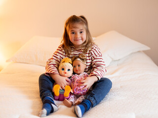 Smiling little blond girl sitting on bed in casual clothes holding two dolls