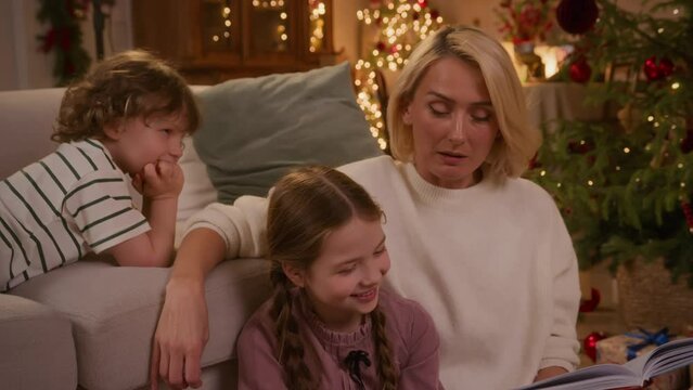 mom, her little daughter and son sit on floor together near Christmas tree at home, and read fairy tale book, kids listen, smile and laugh at funny stories