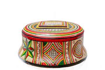 A circular box closed with its lid. With lots of patterns and colors. A checkerboard, curves, triangles, stars and rectangles in beige, yellow, red and green. Studio shot isolated in white background.