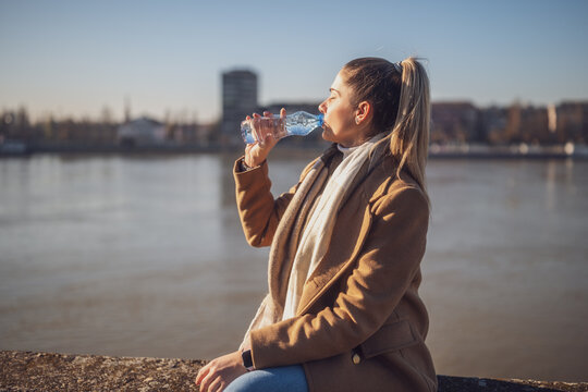 Woman enjoys drinking water while sitting by the river on a sunny winter day.  Toned image.
