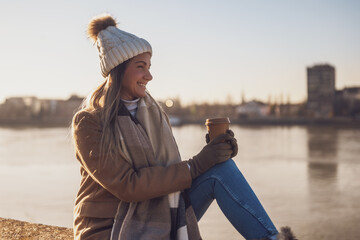 Beautiful woman in warm clothing enjoys  drinking coffee and resting by the river on a sunny winter day.. Toned image.