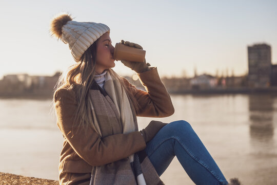 Beautiful woman in warm clothing enjoys  drinking coffee and resting by the river on a sunny winter day.. Toned image.