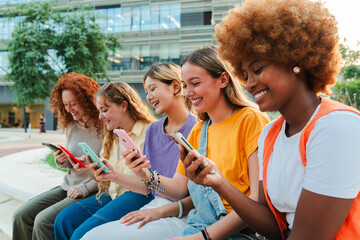 Group of smiling multiracial college students females watching social media app or chatting with their cellphones sitting outside of the university campus. Teenage girls using phones to post photos