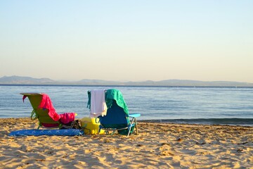 Folding chairs towels and cooler for snacks stand on the beach towards the sea during the sunset,...