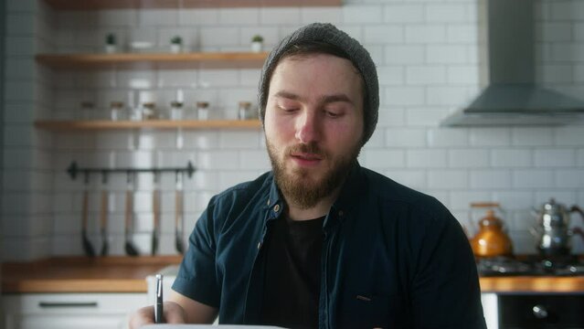 Young angry manager man with beanie sitting in modern kitchen at home, talking, showing reports in video conference call, angry at staff, throwing papers. Webcam view