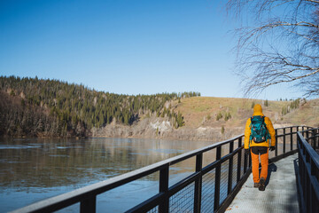 A solo solo trip with a backpack in a mountainous area, a guy walks on a bridge along the river, trekking in cool weather, a sunny day in nature.