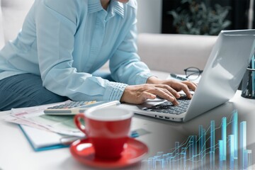 Mujer de negocios vestida con su blusa celeste escribe en su laptop envía mails y whatsapps as su contactos mientras bebe su café en su taza roja. Un gráfico de crecimiento económico al pie de imagen.